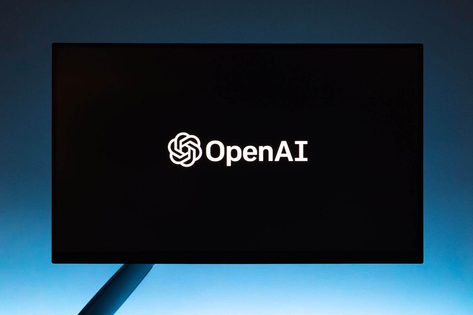 蓝色背景前的黑色显示器，屏幕中央显示白色 OpenAI 标志和文字。真实摄影资料图。
