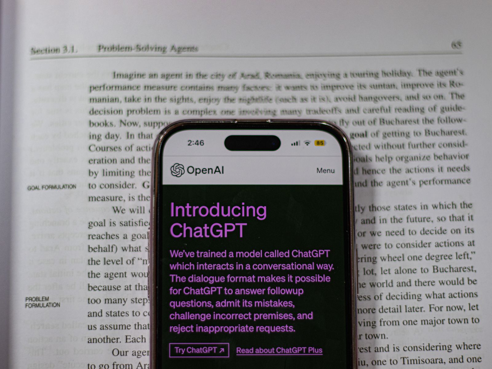 一本打开的英文书上放着手机，手机屏幕显示 OpenAI 的 Introducing ChatGPT 介绍网页。真实摄影资料图。