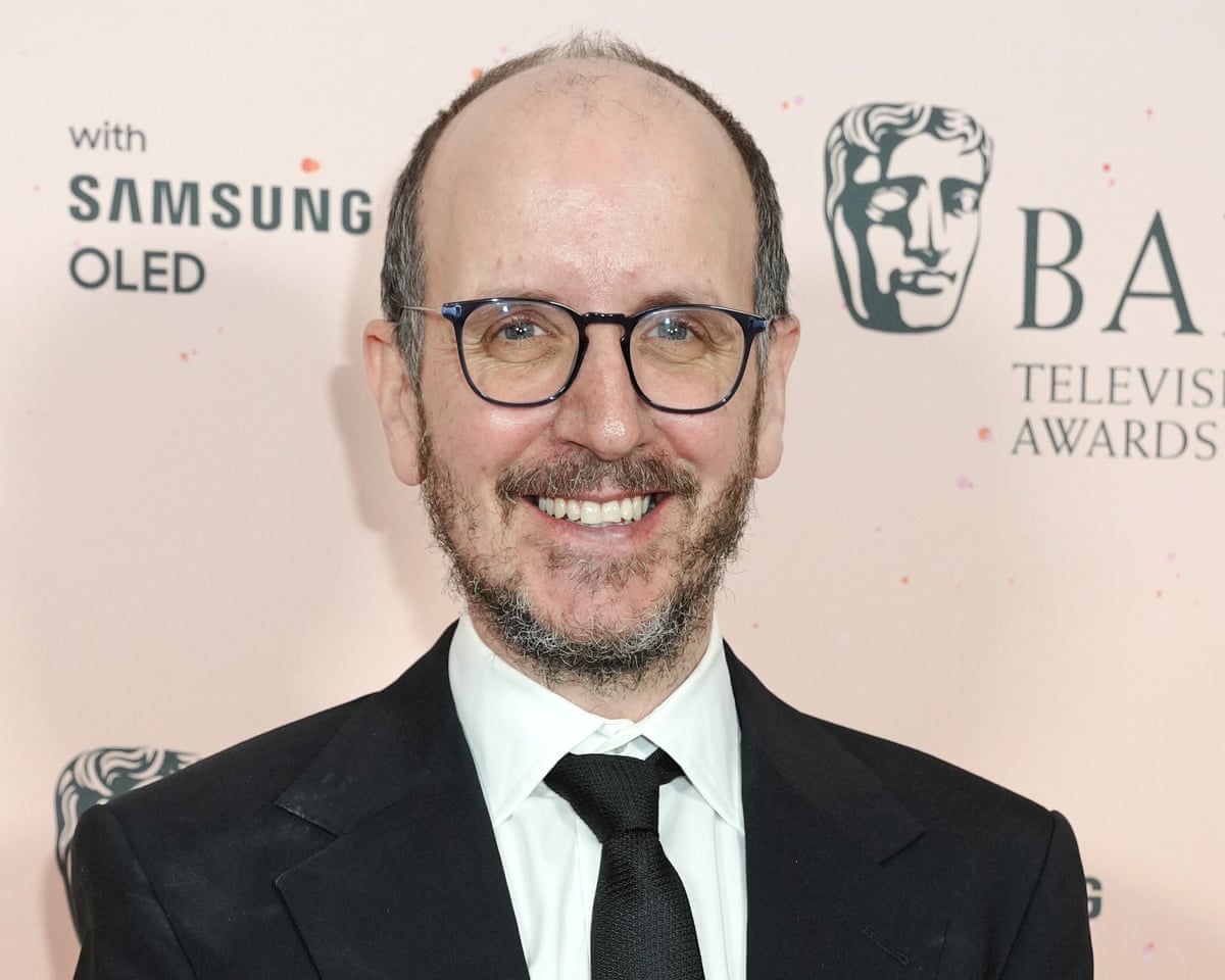 一名戴黑框眼镜、穿黑西装白衬衫的男子在 BAFTA 电视奖背景板前微笑；原报道图注标为编剧 Jack Thorne。